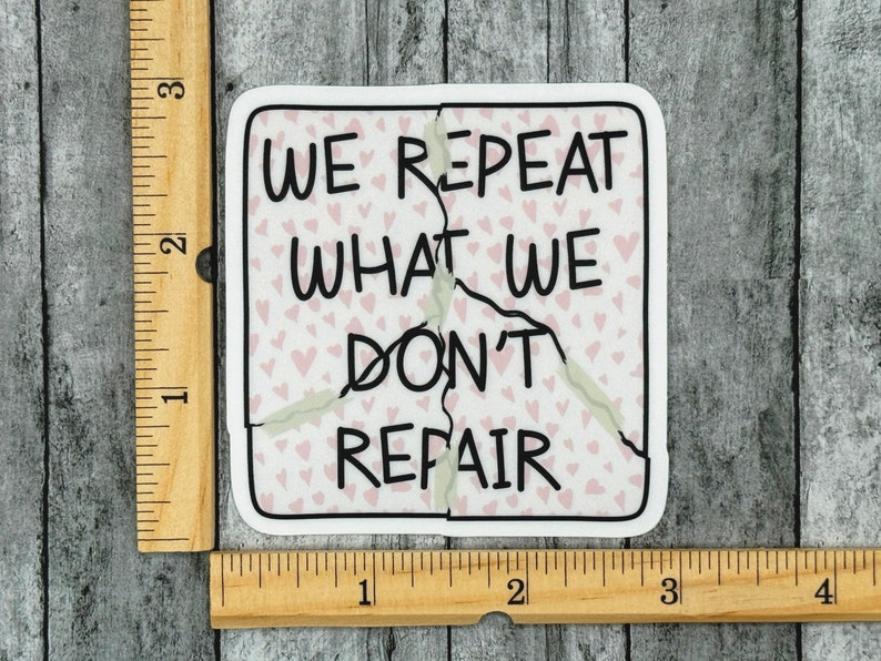 May include: A white sticker with a pink background and a black outline. The sticker is cracked down the middle and says "We repeat what we don't repair" with a pattern of pink hearts in the background.