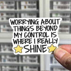 May include: A white die-cut sticker with black text that reads: WORRYING ABOUT THINGS BEYOND MY CONTROL IS WHERE I REALLY SHINE. The sticker features a yellow five-pointed star on each side of the word SHINE, with a subtle speckled texture.