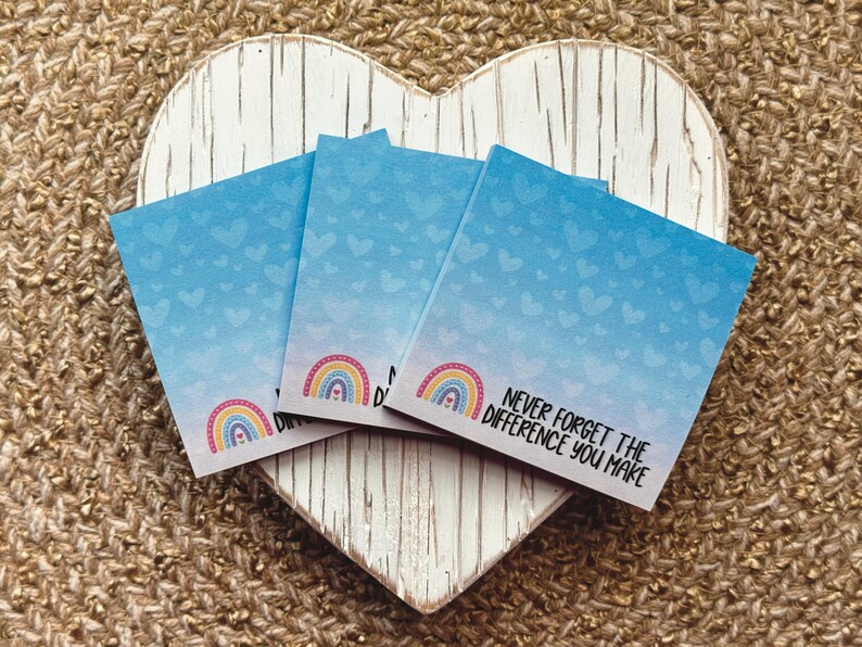 May include: Three square sticky notes with a light blue gradient background featuring a pattern of small hearts. Each note displays a colorful rainbow graphic and the black printed text, "NEVER FORGET THE DIFFERENCE YOU MAKE."