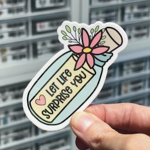May include: A die-cut sticker shaped like a glass bottle with a light blue tint. The bottle contains a yellow label with the black text "Let Life SURPRISE YOU" and a small pink heart. A pink flower with green leaves sits on the bottle neck.