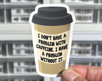May include: A die-cut sticker shaped like a tan coffee cup with a dark gray lid. The cup features a textured sleeve with the black text: I DON'T HAVE A PROBLEM WITH CAFFEINE. I HAVE A PROBLEM WITHOUT IT. The design has a white border outline.