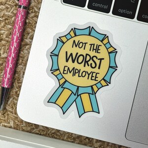May include: A sticker with a yellow and blue ribbon design, featuring the text "NOT THE WORST EMPLOYEE" in black. The sticker is on a laptop, next to a pink pen.