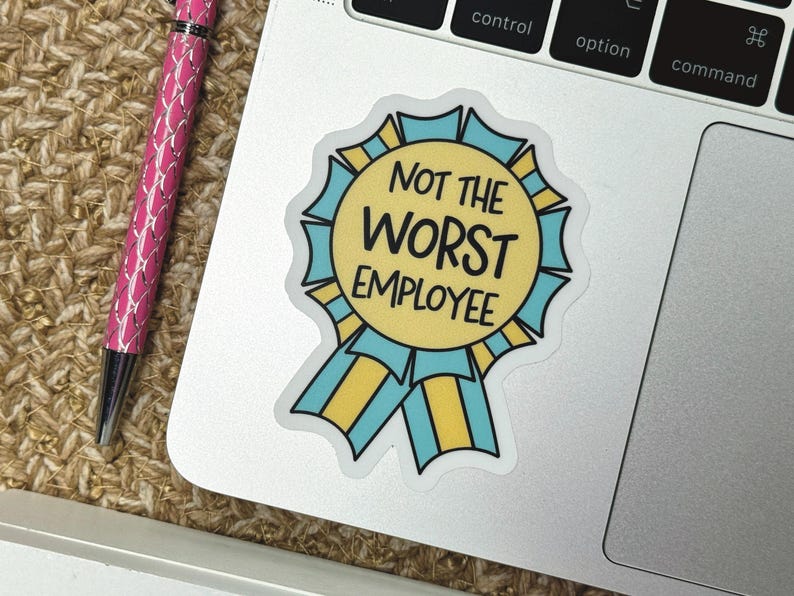 May include: A sticker with a yellow and blue ribbon design, featuring the text "NOT THE WORST EMPLOYEE" in black. The sticker is on a laptop, next to a pink pen.
