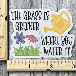 May include: A die-cut sticker featuring a yellow watering can, a purple flower, a pink flower, and green grass with the text "THE GRASS IS GREENER WHERE YOU WATER IT". A wooden ruler shows the sticker is approximately 3 inches wide and 2 inches tall.