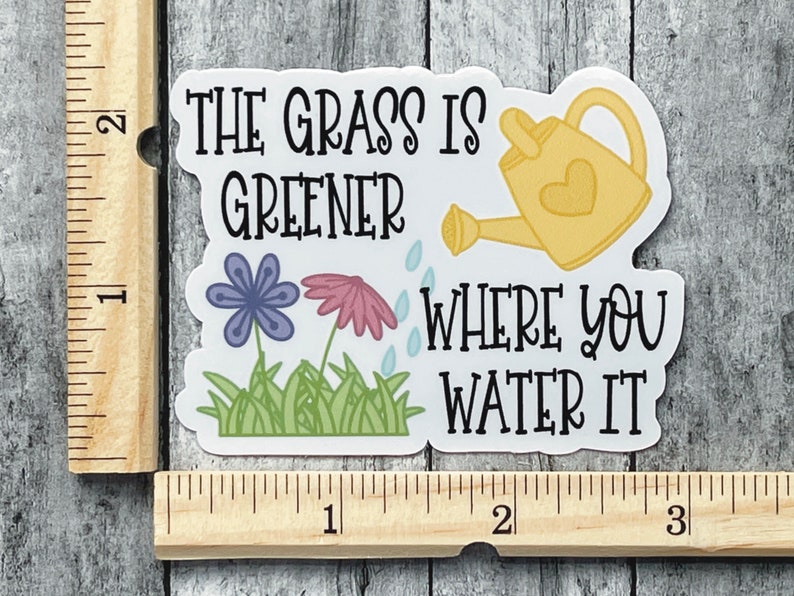 May include: A die-cut sticker featuring a yellow watering can, a purple flower, a pink flower, and green grass with the text "THE GRASS IS GREENER WHERE YOU WATER IT". A wooden ruler shows the sticker is approximately 3 inches wide and 2 inches tall.