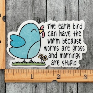 Op de afbeelding: Een gestanste sticker met een lichtblauwe vogel die een roze worm vasthoudt, met de tekst "The early bird can have the worm because worms are gross and mornings are stupid." Een houten liniaal toont een breedte van ongeveer 7,6 cm.