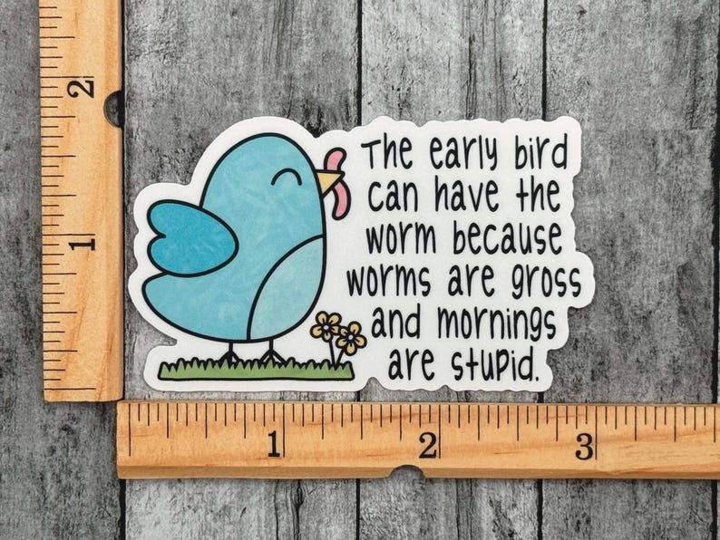 Op de afbeelding: Een gestanste sticker met een lichtblauwe vogel die een roze worm vasthoudt, met de tekst "The early bird can have the worm because worms are gross and mornings are stupid." Een houten liniaal toont een breedte van ongeveer 7,6 cm.