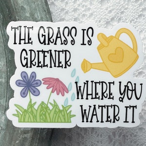 May include: A rectangular sticker featuring the black text THE GRASS IS GREENER WHERE YOU WATER IT, a yellow watering can with a heart, a purple flower, a pink flower, green grass, and light blue water droplets on a white background.