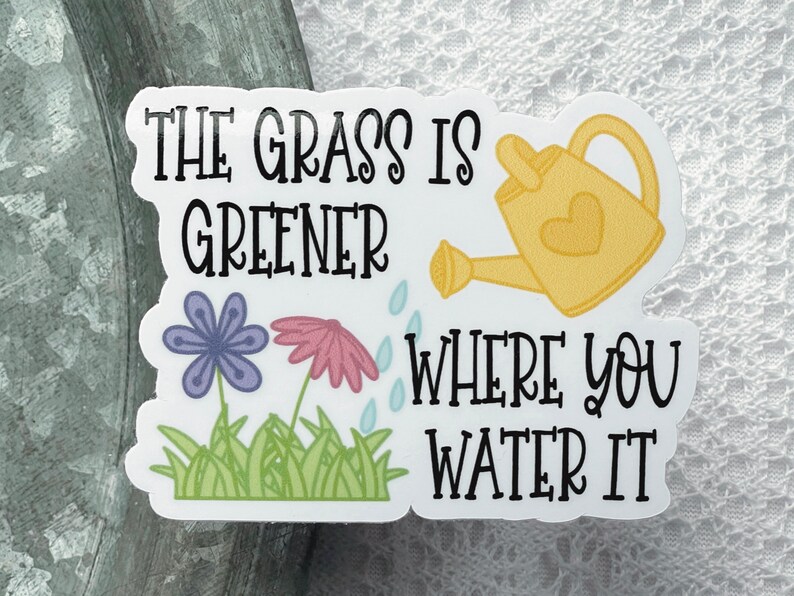 May include: A rectangular sticker featuring the black text THE GRASS IS GREENER WHERE YOU WATER IT, a yellow watering can with a heart, a purple flower, a pink flower, green grass, and light blue water droplets on a white background.