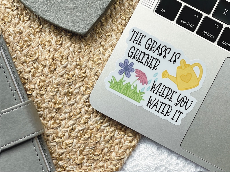 May include: A vinyl sticker featuring a yellow watering can, a purple flower, a pink flower, and green grass. The text reads, THE GRASS IS GREENER WHERE YOU WATER IT. The sticker is applied to the silver surface of a laptop keyboard area.