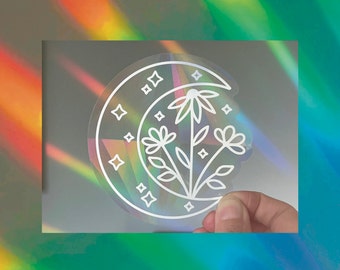 May include: A circular holographic sticker featuring a white line art design of a crescent moon filled with three stylized flowers and several four-pointed stars. The sticker has a semi-transparent finish that reflects rainbow light patterns.