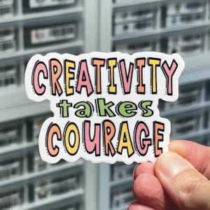 May include: A white sticker with the text "CREATIVITY takes COURAGE" in colorful, outlined letters. The sticker is held in a hand against a blurry background.