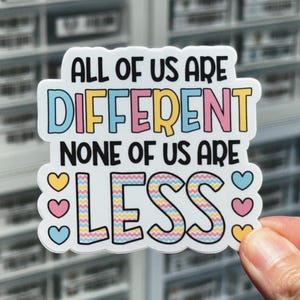 May include: A white sticker with the text "All of us are different, none of us are less." The text is in black and is surrounded by a colorful wavy border. There are three small hearts in each corner of the sticker, in yellow, pink, and blue.