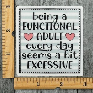 May include: A square sticker with a light blue and white horizontal striped background and a black border. The text reads being a FUNCTIONAL ADULT every day seems a bit EXCESSIVE with two pink hearts. A wooden ruler shows it is 3 inches wide.