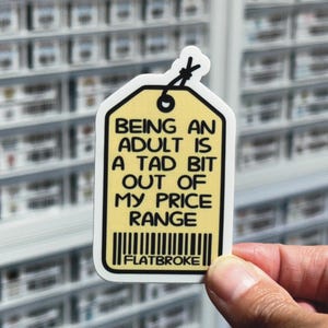 May include: A yellow sticker in the shape of a price tag with the text "BEING AN ADULT IS A TAD BIT OUT OF MY PRICE RANGE" in black. The sticker also has a barcode and the word "FLATBROKE".