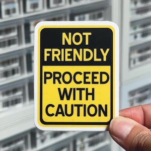 May include: A rectangular sticker with rounded corners, featuring the text "NOT FRIENDLY PROCEED WITH CAUTION" in bold, black letters on a yellow background. The sticker is held by a hand.