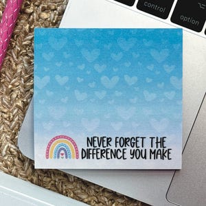May include: A square sticky note with a blue gradient background featuring a pattern of light blue hearts. The bottom left corner displays a colorful rainbow icon, and the text reads, "NEVER FORGET THE DIFFERENCE YOU MAKE" in black.