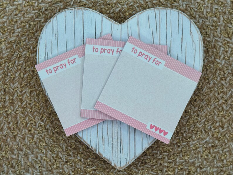 May include: Three square paper note pads with a light pink and white striped border at the top and bottom. The text "to pray for" is printed in pink at the top of each pad. The bottom right corner of the top pad features four small pink hearts.