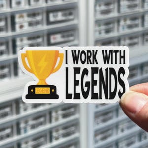 May include: A die-cut sticker featuring a yellow trophy with a lightning bolt icon on the cup and the word LEGEND on the base. To the right of the trophy, the black text reads I WORK WITH LEGENDS in a bold, sans-serif font on a white background.