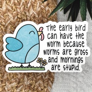Op de afbeelding: Een gestanste sticker met een lichtblauwe cartoonvogel die op groen gras naast kleine gele bloemen staat. De vogel houdt een roze worm in zijn snavel. De zwarte tekst luidt: The early bird can have the worm because worms are gross and mornings are stupid.