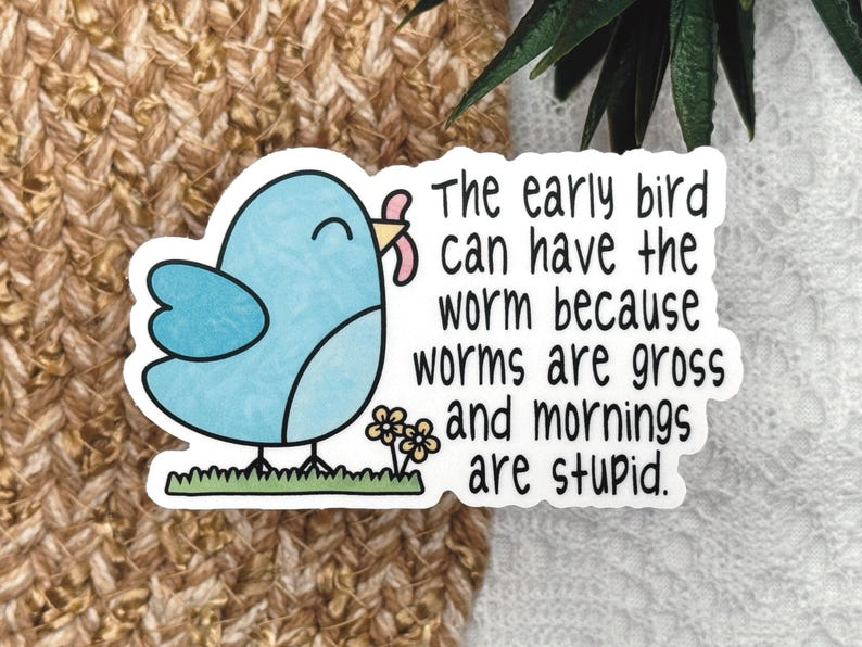 Op de afbeelding: Een gestanste sticker met een lichtblauwe cartoonvogel die op groen gras naast kleine gele bloemen staat. De vogel houdt een roze worm in zijn snavel. De zwarte tekst luidt: The early bird can have the worm because worms are gross and mornings are stupid.