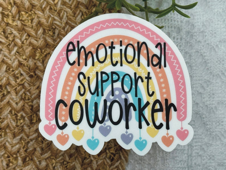 Puede incluir: Una pegatina troquelada con un arco&iacute;ris de colores pastel y el texto negro "emotional SUPPORT COWORKER" impreso en el centro. El dise&ntilde;o del arco&iacute;ris incluye peque&ntilde;as formas de coraz&oacute;n colgantes en rosa, amarillo, azul y morado en la base.