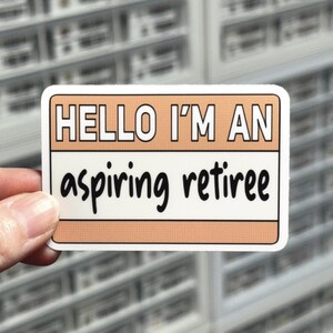 May include: A rectangular sticker with a peach background and white border. The text "HELLO I'M AN" is at the top, and "aspiring retiree" is written below. The sticker is held by a person's fingers.