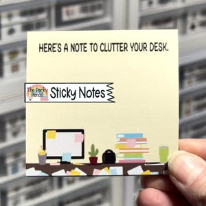 May include: A square, pale yellow sticky note featuring the black text "HERE'S A NOTE TO CLUTTER YOUR DESK." at the top. The bottom displays a colorful illustration of a desk with a computer, a cactus, a stack of books, and scattered papers.