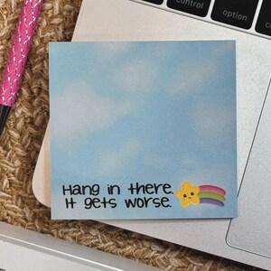 May include: A square sticky note with a light blue sky and white cloud background. The text reads "Hang in there. It gets worse." next to a small yellow star character with a sad face and a colorful rainbow trail extending to the right.