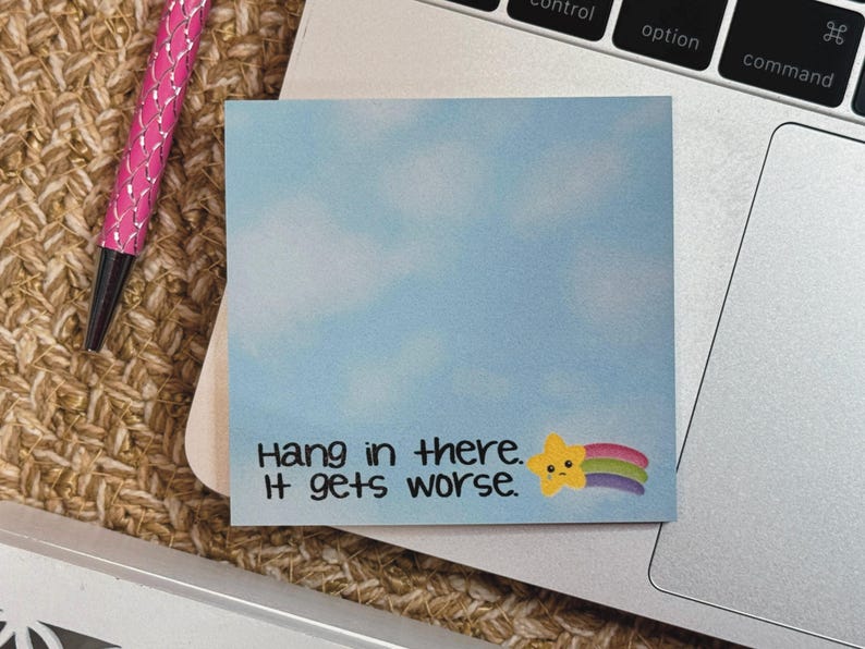 May include: A square sticky note with a light blue sky and white cloud background. The text reads "Hang in there. It gets worse." next to a small yellow star character with a sad face and a colorful rainbow trail extending to the right.