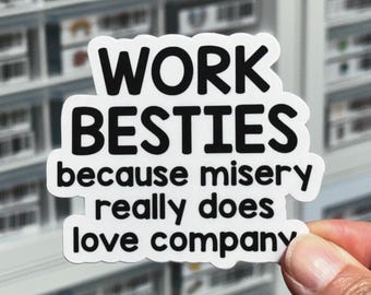 May include: A white sticker with black text that reads "WORK BESTIES because misery really does love company." The sticker is held by a hand in front of a blurred background of shelves.