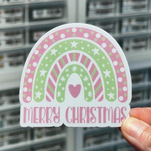 May include: A pink and green rainbow sticker with white stars and snowflakes. The rainbow has a pink heart in the center and the text "Merry Christmas" at the bottom.