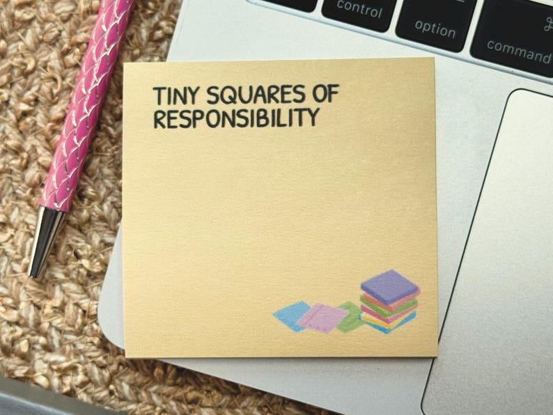 Peut inclure: Un bloc-notes dor&eacute; avec le texte "TINY SQUARES OF RESPONSIBILITY" en noir. La note comporte une pile de carr&eacute;s color&eacute;s en bas &agrave; droite. Un stylo rose avec une pointe argent&eacute;e est &agrave; gauche.