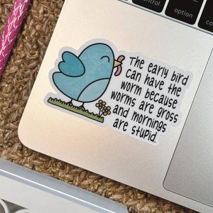 Op de afbeelding: Een gestanste sticker met een lichtblauwe vogel die een roze worm in zijn snavel houdt, staand op een strook groen gras met kleine gele bloemetjes. De tekst luidt: The early bird can have the worm because worms are gross and mornings are stupid.