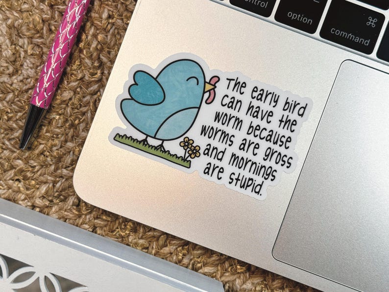 Op de afbeelding: Een gestanste sticker met een lichtblauwe vogel die een roze worm in zijn snavel houdt, staand op een strook groen gras met kleine gele bloemetjes. De tekst luidt: The early bird can have the worm because worms are gross and mornings are stupid.