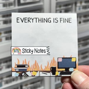 May include: White sticky note with the text "EVERYTHING IS FINE" in black. The bottom of the note features a cartoon illustration of a computer, books, and a coffee mug, with flames rising up. The words "Sticky Notes" are also on the note.