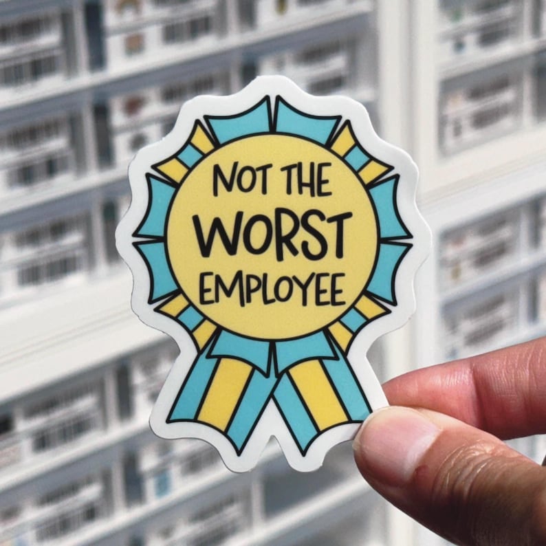 May include: A sticker with the text "NOT THE WORST EMPLOYEE" in black letters. The sticker is shaped like a yellow and blue award ribbon. The background is a blurred image of shelves.