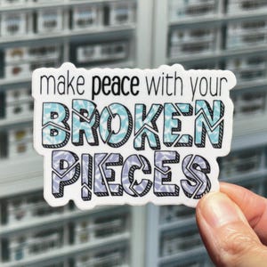 May include: White sticker with the text "make peace with your broken pieces" in black. The words are outlined in black and filled with a blue and purple geometric pattern.