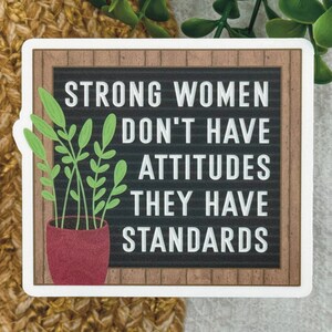 May include: A square sticker featuring a black letter board with a brown wood-look frame. The white text reads: STRONG WOMEN DON'T HAVE ATTITUDES THEY HAVE STANDARDS. A potted green plant with a red pot is positioned on the left side of the board.