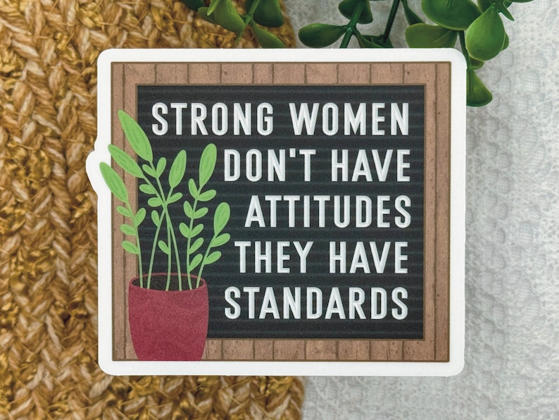 May include: A square sticker featuring a black letter board with a brown wood-look frame. The white text reads: STRONG WOMEN DON'T HAVE ATTITUDES THEY HAVE STANDARDS. A potted green plant with a red pot is positioned on the left side of the board.