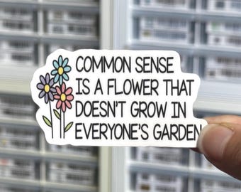 May include: A white die-cut sticker featuring three illustrated flowers in blue, purple, and pink with green stems. The black text reads: COMMON SENSE IS A FLOWER THAT DOESN'T GROW IN EVERYONE'S GARDEN. The sticker has a clean, white border.