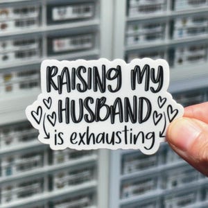May include: White sticker with black text that reads "Raising my husband is exhausting" with hearts and arrows around the text.