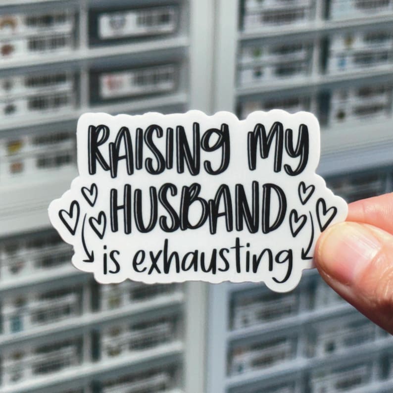 May include: White sticker with black text that reads "Raising my husband is exhausting" with hearts and arrows around the text.