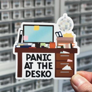 May include: A die-cut sticker featuring a brown desk with a computer monitor, a disco ball, and office supplies. The text "PANIC AT THE DESKO" is printed in bold black letters on the front of the desk, which includes three drawers and various papers.