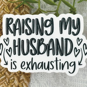 May include: White sticker with black text that reads "Raising my husband is exhausting" with a series of black hearts around the word "husband".
