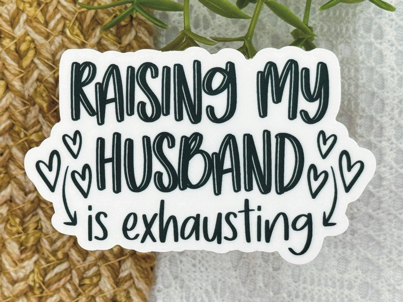 May include: White sticker with black text that reads "Raising my husband is exhausting" with a series of black hearts around the word "husband".
