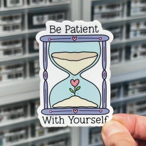 May include: A blue and purple hourglass sticker with a pink heart growing inside. The sticker says "Be Patient With Yourself".