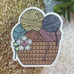 May include: A brown wicker basket filled with colorful yarn balls. The yarn balls are blue, yellow, pink, and brown. There are white buttons and a pink heart on the front of the basket.