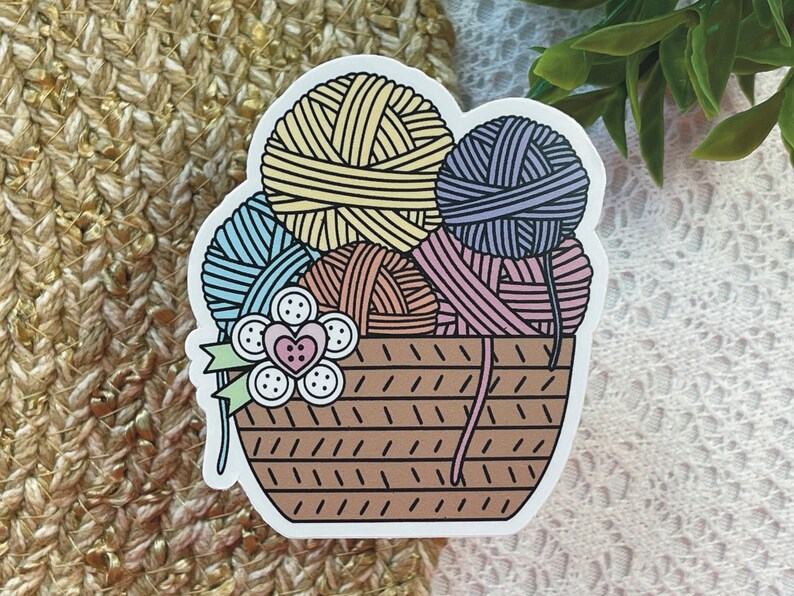 May include: A brown wicker basket filled with colorful yarn balls. The yarn balls are blue, yellow, pink, and brown. There are white buttons and a pink heart on the front of the basket.