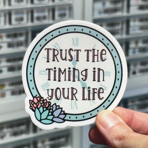 May include: A round sticker featuring a clock face design with a light blue border and Roman numerals. The text "TRUST THE TIMING IN YOUR LIFE" is printed in dark brown letters. A cluster of pink, blue, and purple succulent illustrations sits at the bottom.